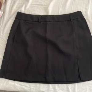 Svart kortkjol med slits - Snygg svart kortkjol med en diskret slits på sidan. Kjolen har en klassisk design med hällor för bälte och en stilren look. Perfekt för att matcha med olika toppar och accessoarer.