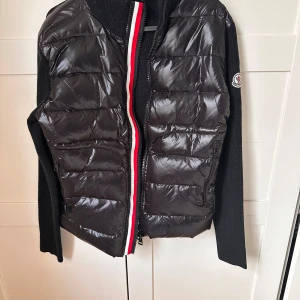 Svart pufferjacka från Moncler - Snygg svart pufferjacka från Moncler med stickade ärmar och en cool dragkedja i rött, vitt och svart. Jackan har ett quiltat mönster och Moncler-loggan på ärmen. Perfekt för kyligare dagar när du vill hålla stilen.