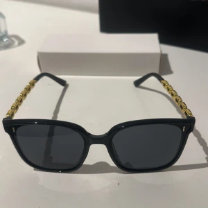 Svarta solglasögon med gulddetaljer - Snygga svarta solglasögon med fyrkantig form och coola guldfärgade kedjedetaljer på skalmarna. Perfekta för att ge en lyxig touch till din stil. De mörka glasen ger ett stilrent intryck och skyddar mot solen.