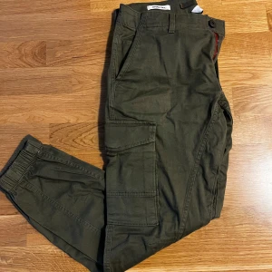 Gröna cargobyxor från Jack & Jones - Snygga gröna cargobyxor från Jack & Jones med flera praktiska fickor. Byxorna har en tapered passform och är perfekta för en avslappnad stil. De har knappar och dragkedja i gylfen. Storlek 29/30 och dom är i bra skick 