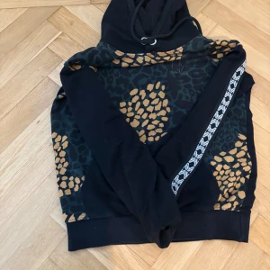 Marcel von berlin hoodie - Säljer en cool svart hoodie med leopardmönster i orange och grönt. Den har en justerbar huva med snörning och en dekorativ rand med mönster längs ärmarna. Perfekt för en avslappnad stil.