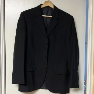 Svart kavaj passar M (Junex, Blue Best) - Elegant svart kavaj med klassisk design och tre knappar framtill. Perfekt för formella tillfällen. Kavajen har fickor med lock och knappar vid ärmsluten.