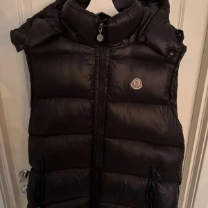 Svart dunväst från Moncler - Snygg svart dunväst från Moncler med dragkedja och hög krage. Perfekt för kyligare dagar och ger en stilren look. Västen har en ikonisk Moncler-logga på bröstet och en unik serietidningsetikett på insidan. Storlek 4 (M/L). Scan kod finns