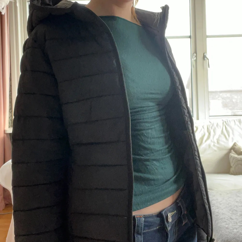 Snygg svart dunjacka med huva och dragkedja framtill. Jackan har en quiltad design som ger en stilren look. Perfekt som vår och höstjacka, kan även funka vid mildare vinter. Lite sliten vid bröstet och ett hål på armbågen.. Takit.