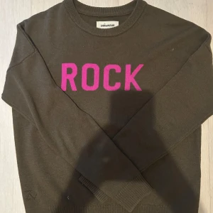 Olivgrön tröja från Zadig & Voltaire - Snygg olivgrön tröja från Zadig & Voltaire med texten 'ROCK' i rosa på bröstet. Tröjan har en rund halsringning och långa ärmar, perfekt för en cool och avslappnad stil. *TRYCK INTE PÅ KÖP NU*