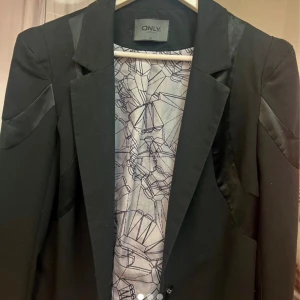 Svart kavaj från ONLY - Snygg svart kavaj från ONLY med stilrena detaljer i satin på axlar och rygg. Perfekt för en chic look. Kavajen har en klassisk krage och är designad för att ge en elegant siluett.