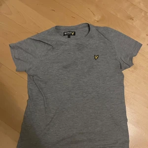 Grå t-shirt från Lyle & Scott - Säljer en stilren grå t-shirt från Lyle & Scott med korta ärmar. Den har en broderad gul logga på bröstet. Perfekt för en avslappnad look.