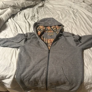 Grå huvtröja från Burberry - Säljer en snygg grå unik huvtröja från Burberry med klassiskt rutigt foder. Tröjan har dragkedja framtill och en bekväm huva. Perfekt för en stilren look med en touch av lyx. 🧥✨den är storlek m passar s skicket är 9/10 inga defekter 