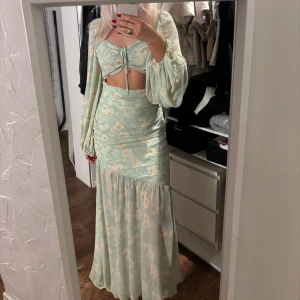 Grön blommig klänning från Adoore - Säljer en vacker grön långklänning från Adoore med blommigt mönster. Klänningen har långa ärmar och en snygg knytning framtill. Perfekt för sommarens festligheter!