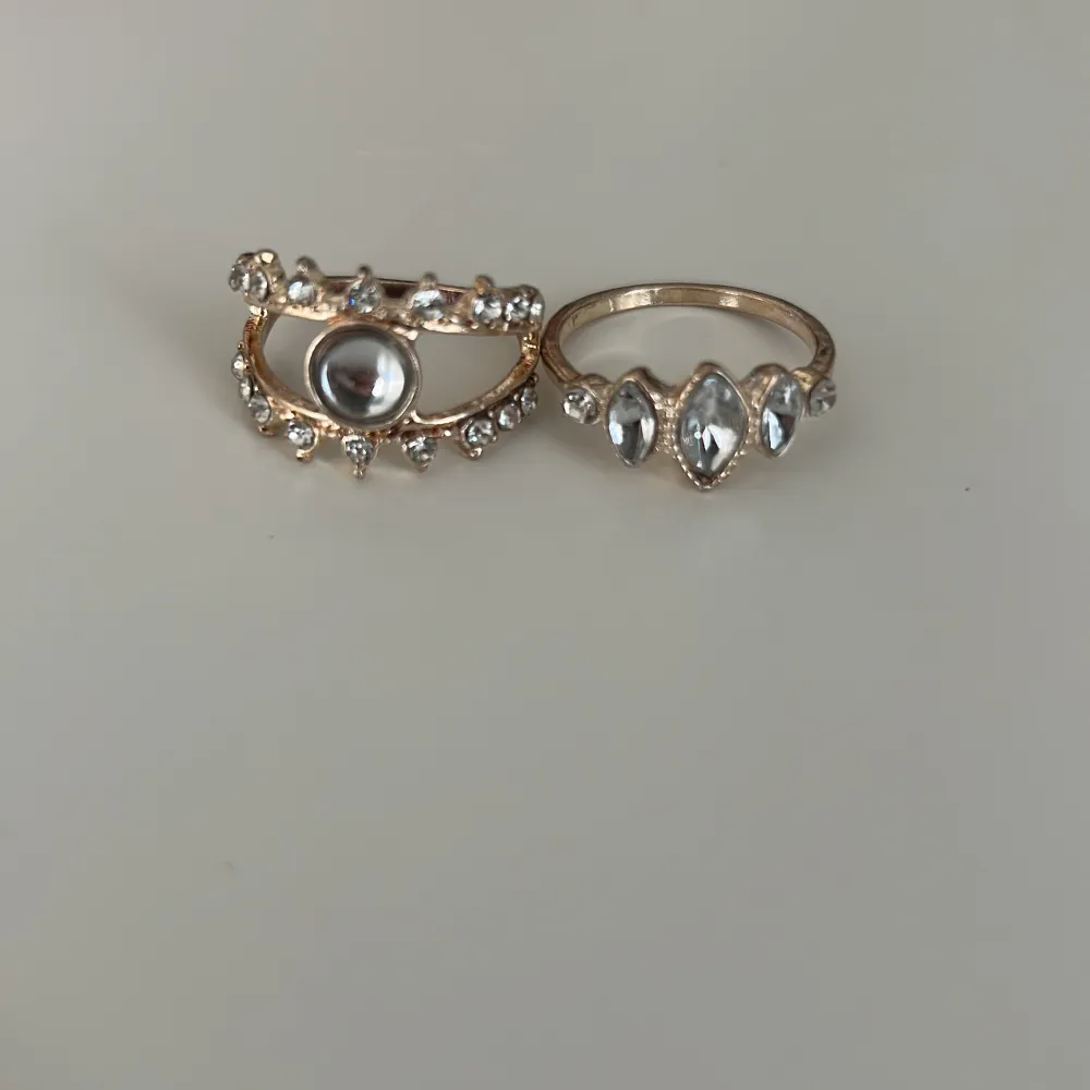 Säljer två stiliga ringar i guldton med glittrande stenar. Den ena ringen har en stor central sten omgiven av mindre stenar, medan den andra har tre större stenar i rad. Perfekta för att ge en touch av glamour till din look ❤️ (#85). Asusteet.