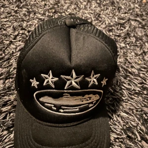 Svart keps med broderade stjärnor - Snygg svart keps med broderade stjärnor och ett motiv framtill. Kepsen har ett nätmaterial på sidorna och justerbar passform baktill. Perfekt för en cool och avslappnad stil.