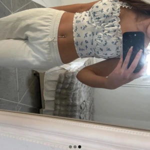 Vit blommig linnetopp - Söt vit linnetopp med blå blommor och volangdetaljer på axelbanden. Toppen har en ribbad struktur och knytning framtill för en extra touch. Perfekt för en somrig look!