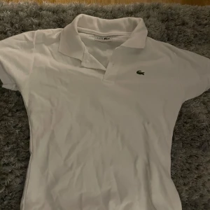 Vit pikétröja från Lacoste - Säljer en klassisk vit pikétröja från Lacoste med den ikoniska krokodilloggan på bröstet. Tröjan har korta ärmar och en stilren krage. Perfekt för en avslappnad och stilren look.