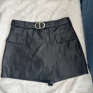 Svart skinnbyxkjol med bälte - Snygg svart fejkskinnbyxkjol med ett elegant bälte och guldfärgat spänne. Den har en omlott-design som ger en modern look. Perfekt att matcha med en stickad tröja på kallare dagar eller med ett linne på varmare dagar!💕💕🌸🌸