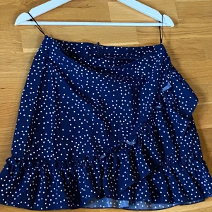 Blå prickig kjol med volang!  - Säljer en söt blå kortkjol med vita prickar och volangdetaljer. Kjolen har en omlott-design som ger en fin passform. Perfekt för sommarens alla tillfällen! Dragkedja bak. Totala längd 42 cm. 