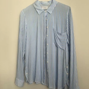 Blå och vit randig skjorta från Pull&Bear - Snygg blå och vit randig skjorta från Pull&Bear i storlek M. Skjortan har långa ärmar och en klassisk krage. Perfekt för en stilren look med knappar framtill och en bröstficka. Liten fläck (se bild).