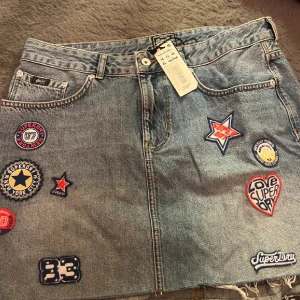 Jeanskjol med patches från Superdry - Cool jeanskjol från Superdry med flera färgglada patches som ger en unik look. Kjolen har en klassisk denimfärg och är perfekt för en avslappnad stil. Den har en knapp och dragkedja framtill samt bälteshällor.