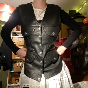 Svart skinnväst från Jennifer - Äkta läder från Jennifer med knappar framtill och fickor. Har en justerbar rem på baksidan. Baksidan är i ett satängliknande material. Oanvänd! 