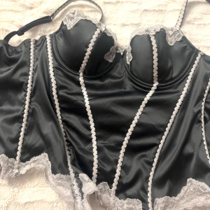Svart satin korsett med spetsdetaljer - Elegant svart korsett i satin med vackra vita spetsdetaljer längs kanterna och kuporna. Den har justerbara axelband och hakknäppning bak för perfekt passform. Perfekt för en stilfull look. Köpt vintage och passar storlek S. 