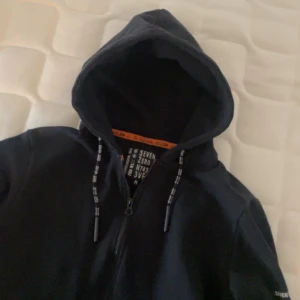 Svart hoodie från Seven Zero - Snygg svart hoodie från Seven Zero med dragkedja och justerbara snören i huvan. Den har tryck på ärmen och bröstet samt praktiska fickor med dragkedja. Perfekt för en avslappnad stil.