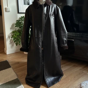 Brun skinnrock från Armani Jeans - Säljer en sjukt snygg brun skinnkappa från Armani Jeans med mjukt foder och knappar framtill. Rocken har en klassisk krage och en bröstficka. Perfekt för kyligare dagar. Ger verkligen mob wife vibes, så iconic💄🖤den är till knäna på mig som är 1,80