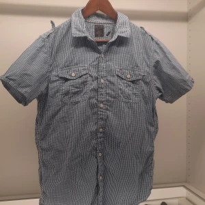 Blå och vit rutig skjorta från Tommy Hilfiger - Snygg blå och vit rutig skjorta från Tommy Hilfiger i storlek medium. Skjortan har korta ärmar och knappar framtill samt två bröstfickor med knappstängning. Perfekt för en avslappnad stil med en touch av klassisk design.