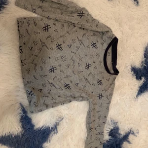 Grå långärmad tröja med mönster från Lupilu - Säljer en grå långärmad tröja från Lupilu med ett coolt mönster av symboler som stjärnor, blixtar och hashtags. Perfekt för vardagsbruk och lek. Tröjan har en mörkare kant vid halsen som ger en snygg kontrast.