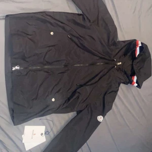 Svart vindjacka från Moncler - Snygg svart vindjacka från Moncler med dragkedja och huva. Jackan har en cool randig detalj i rött, vitt och blått på huvan samt en logotyp på ärmen. Perfekt för blåsiga dagar!