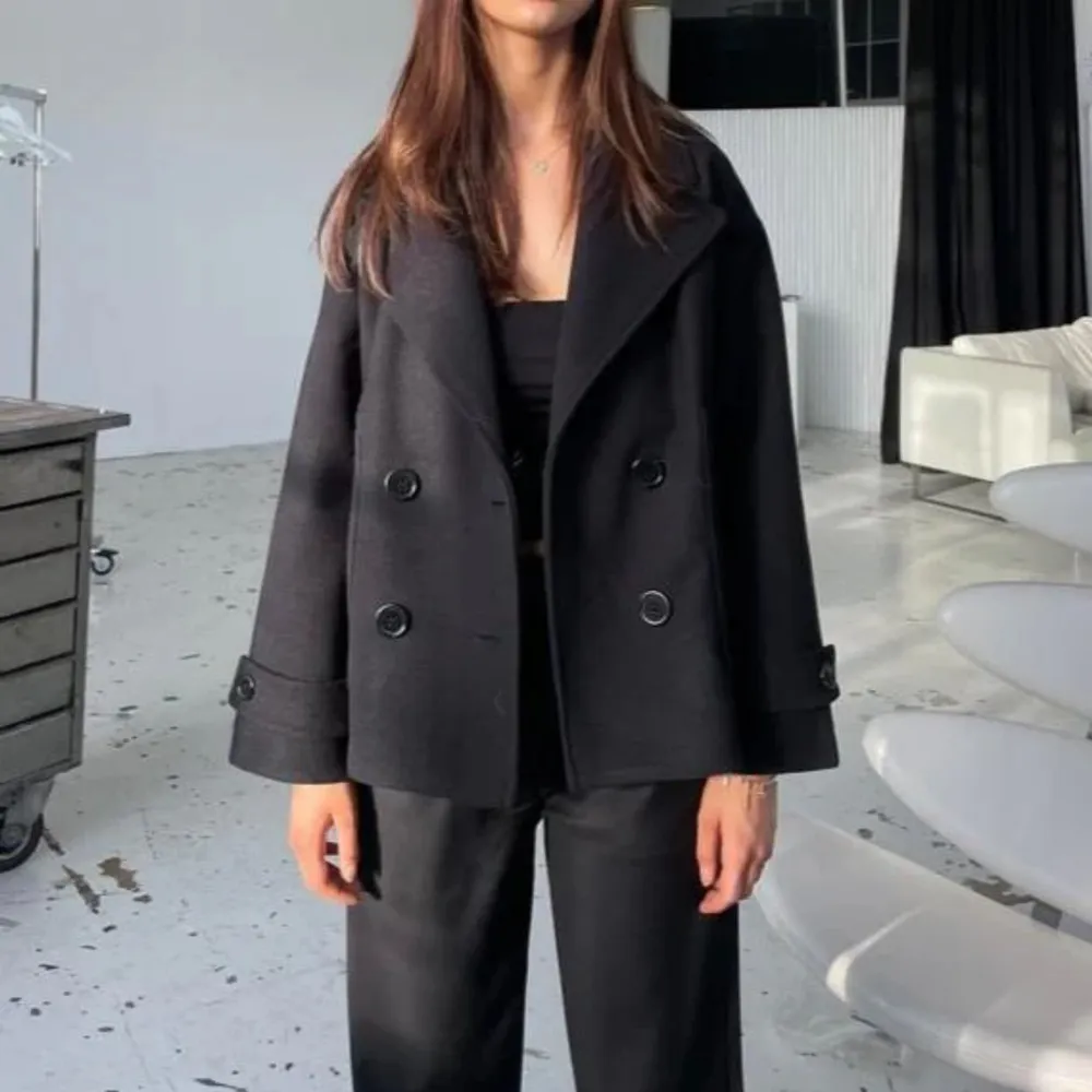Snygg svart kappa med dubbelknäppning och bred krage. Jackan har en kortare passform och långa ärmar med dekorativa knappar vid ärmsluten. Perfekt för en stilren look. Från design by si, svart. Takit.