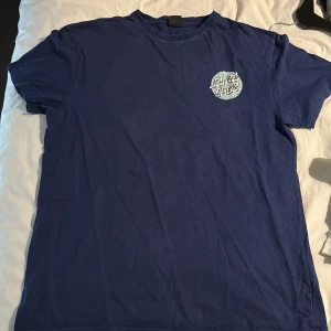 Blå t-shirt från Santa Cruz - Säljer en blå t-shirt från Santa Cruz med ett coolt tryck på bröstet. Perfekt för en avslappnad stil. T-shirten har korta ärmar och en rund halsringning.