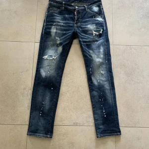 Dsquared2 jeans med slitningar Bella Ciao - Snygga mörkblå jeans med coola slitningar och färgstänk. De har en klassisk femficksdesign och en knappgylf. Perfekta för en avslappnad stil med en edgy touch. Säljes då jag ej använder dem någon mer. Storlek 48, sitter som w30-32. L30-32. Slim fit med skön passform. Kommer ej ihåg vilken style det var. 