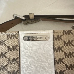 Axelväska från Michael Kors - Snygg axelväska från Michael Kors med det klassiska MK-mönstret i beige och brunt. Väskan har en vit panel i mitten och en justerbar axelrem i brunt läder. Perfekt för att ge din outfit en lyxig touch.