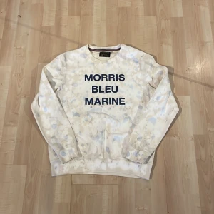 Morris tröja med snyggt mönster - Säljer en vit sweatshirt från Morris med texten 'Morris Bleu Marine' i blått. Tröjan har ett unikt mönster med ljusa fläckar och ribbade muddar vid ärmslut och nederkant. Perfekt för en avslappnad stil.
