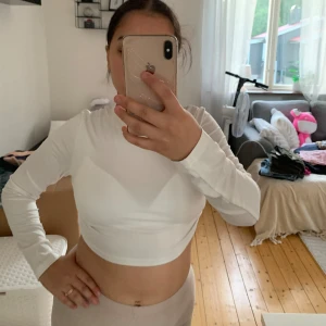 Vit långärmad croppad topp med öppen rygg - Säljer en snygg vit långärmad croppad topp med öppen rygg och knytband. Perfekt för både vardag och fest! Toppen är i ett stretchigt material som sitter tight och bekvämt på kroppen. Den har en stilren design framtill och en cool detalj med öppen rygg som knyts ihop med band.