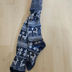 Mönstrade stickade knästrumpor - Säljer ett par supermysiga stickade strumpor med ett fint mönster i blått och vitt. Perfekta för vintern och håller fötterna varma och sköna. Strumporna har ett snyggt mönster med renar och snöflingor, vilket gör dem extra festliga för julen.100% ull