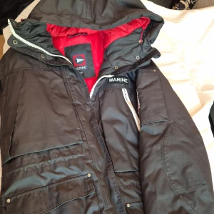 Svart vinterjacka från Marine - Säljer en svart vinterjacka från Marine i storlek XL. Jackan har en röd insida och är utrustad med en dragkedja och knappar. Den har en huva och flera fickor för extra förvaring. Perfekt för kalla vinterdagar. Observera att dragkedjan är något sliten, se bild.