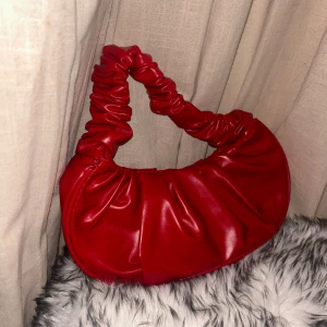 Röd axelväska i syntetmaterial - Säljer en superfin röd axelväska med en rynkad design på handtaget och en enkel knappstängning. Perfekt för att piffa upp vilken outfit som helst! 💖