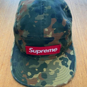 Camouflage keps från Supreme - Säljer en snygg camouflage keps från Supreme. Kepsen har ett klassiskt camo-mönster i gröna, bruna och svarta nyanser. Den har en röd Supreme-logga på framsidan och är tillverkad i bomull. Perfekt för både vardag och utomhusaktiviteter. Kepsen är i bra skick och redo för en ny ägare!