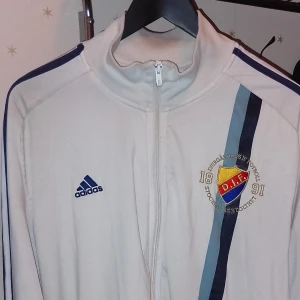 Vit träningsjacka från Adidas med DIF-logga - Säljer en vit träningsjacka från Adidas med Djurgårdens IF-logga på bröstet. Jackan har en dragkedja framtill och blå detaljer på axlarna och bröstet. Perfekt för träning eller att visa ditt stöd för laget!