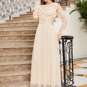 Champagnefärgad långklänning med spetsdetaljer - Säljer en elegant champagnefärgad långklänning med vackra spetsdetaljer och blommönster på överdelen. Klänningen har långa ärmar och en flytande kjol i ett lätt och luftigt material. Perfekt för festliga tillfällen eller en romantisk kväll. 💫