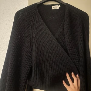 Svart omlottkofta från NA-KD - Säljer en snygg svart omlottkofta från NA-KD. Den är stickad och har en skön passform som passar perfekt för höst och vinter. Koftan har långa ärmar och en stilren design som gör den lätt att matcha med olika outfits. Perfekt för både vardag och fest!
