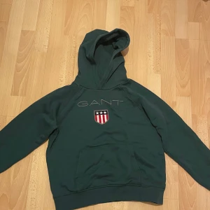 Mörkgrön hoodie från GANT - Säljer en snygg mörkgrön hoodie från GANT. Den har en klassisk design med GANT-loggan och ett coolt emblem på bröstet. Perfekt för en avslappnad stil och passar bra till både jeans och joggers. Superbekväm och perfekt för kyligare dagar!
