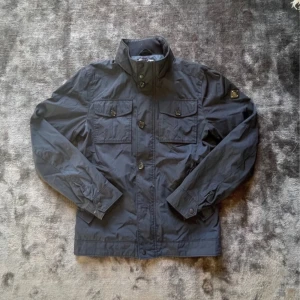 J Lindberg field jacka  - Säljer en jätte snygg J Lindeberg jacka i storlek S. Den är i väldigt bra skick med en gömd luva i kragen. Enda defekten att nämna är att på på armen fattas de en knapp men de syns inte då den täcks över som ni ser i dem två sista bilderna.  