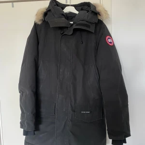 Svart vinterjacka med pälsdetaljer - Säljer en svart vinterjacka från Canada Goose. Den har en mysig pälsdetalj på huvan och är perfekt för kalla vinterdagar. Jackan har långa ärmar och en dragkedja framtill. Den är i bra skick, men har ett litet hål som inte är synligt. Kvitto finns.