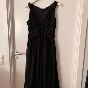 Svart klänning från Lindex - Säljer en elegant svart klänning från Lindex i storlek 38. Klänningen är ärmlös och har en vacker knytning framtill som ger en fin detalj. Materialet känns mjukt och faller snyggt, perfekt för festliga tillfällen. Klänningen går till under knäna