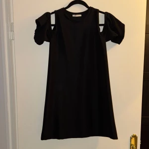 Svart klänning från Zara Trafaluc - Säljer en elegant svart klänning från Zara. Den har en unik design med öppna axlar och puffiga kortärmar som ger en trendig look. Perfekt för både fest och vardag. Materialet känns mjukt och bekvämt mot huden. Passar perfekt för vår och somma