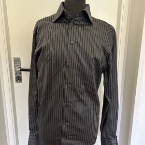 Svart och grå randig skjorta - Säljer en stilren svart och grå randig skjorta med långa ärmar. Skjortan är perfekt för både formella och avslappnade tillfällen. Materialet känns bekvämt och passar bra till hösten och vintern 
