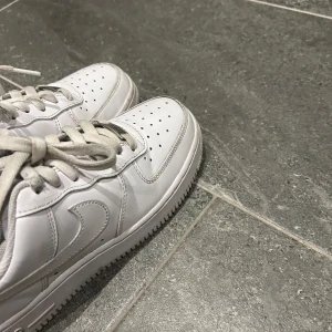 Vita Nike Air Force sneakers - Klassiska vita Nike Air Force sneakers med snörning. Perfekta för en stilren look och passar till det mesta. Skorna har en ikonisk design med perforerade detaljer på tån för extra ventilation. Skorna är som nya men har haft på mig dom få gånger men har inga repor elr skador som ny, nypris 1200kr 