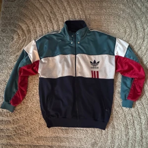  Adidas trackjacket - vintage/Retro (M-L) - Säljer en Snygg vintage| retro Adidas träningsjacka i färgglada block. Jackan har en dragkedja framtill och detaljer i rött, vitt och blått. Designen är typisk för 80- eller 90-talet med Adidas logotyp fram och bak. Jackan är i gott skick, den saknar dock hållaren till dragkedjan men går lätt att ersätta med en ny!  Perfekt för dig som uppskattar vintagemode. Storlek: F180 (M-L) 
