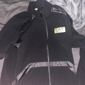 Svart tjocktröja från Emporio Armani - Snygg svart tjocktröja från Emporio Armani med dragkedja och EA7-logga på bröstet. Tjocktröja har en stilren design med långa ärmar och detaljer i syntetmaterial. Perfekt för en sportig look.